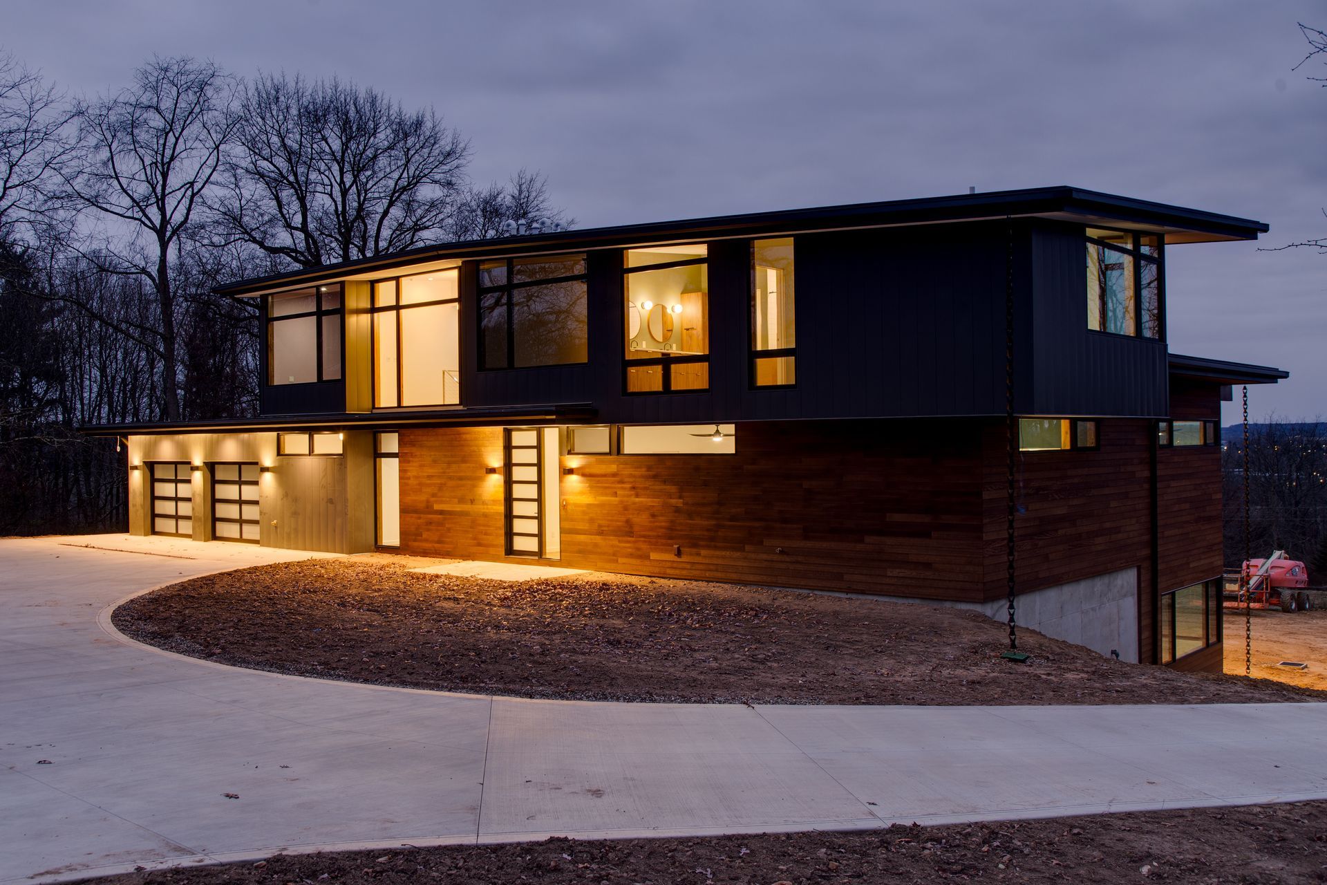 Modern two-story house with warm interior lights at dusk, curved driveway, and bare trees outside