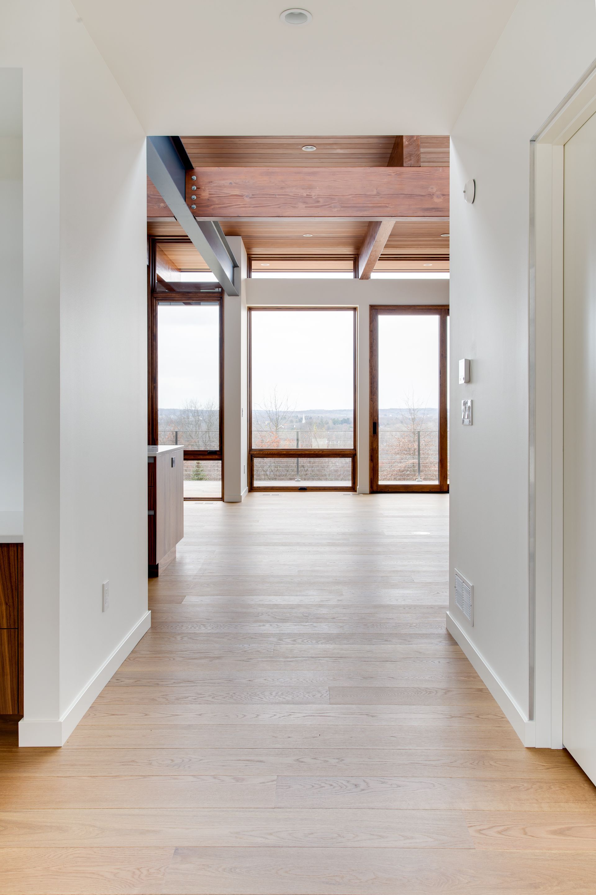 Bright hallway with light wood floors leading to large windows and a balcony, exposed wood ceiling above