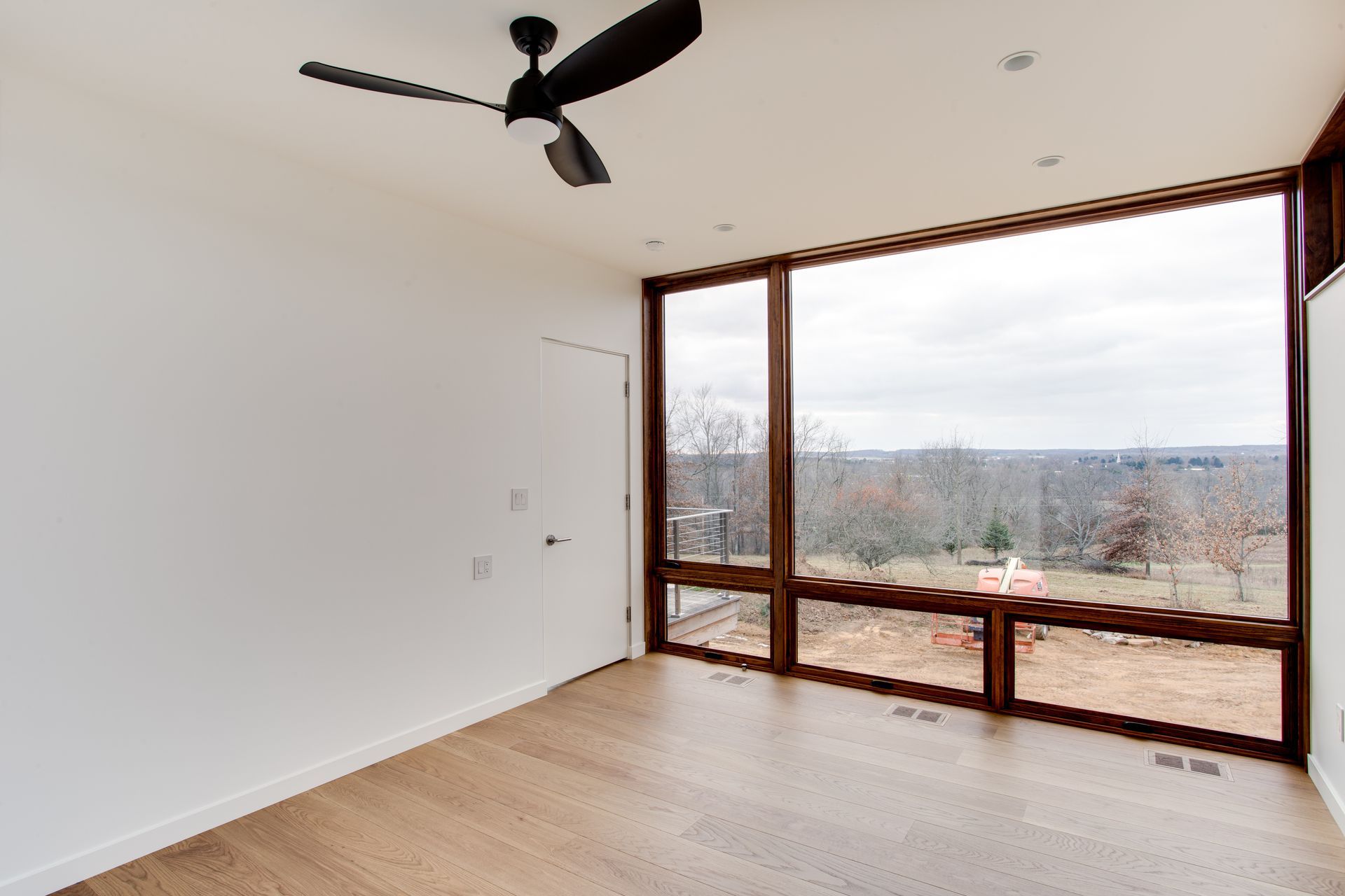 Empty room with white walls, wood floor, ceiling fan, and large windows overlooking a city view
