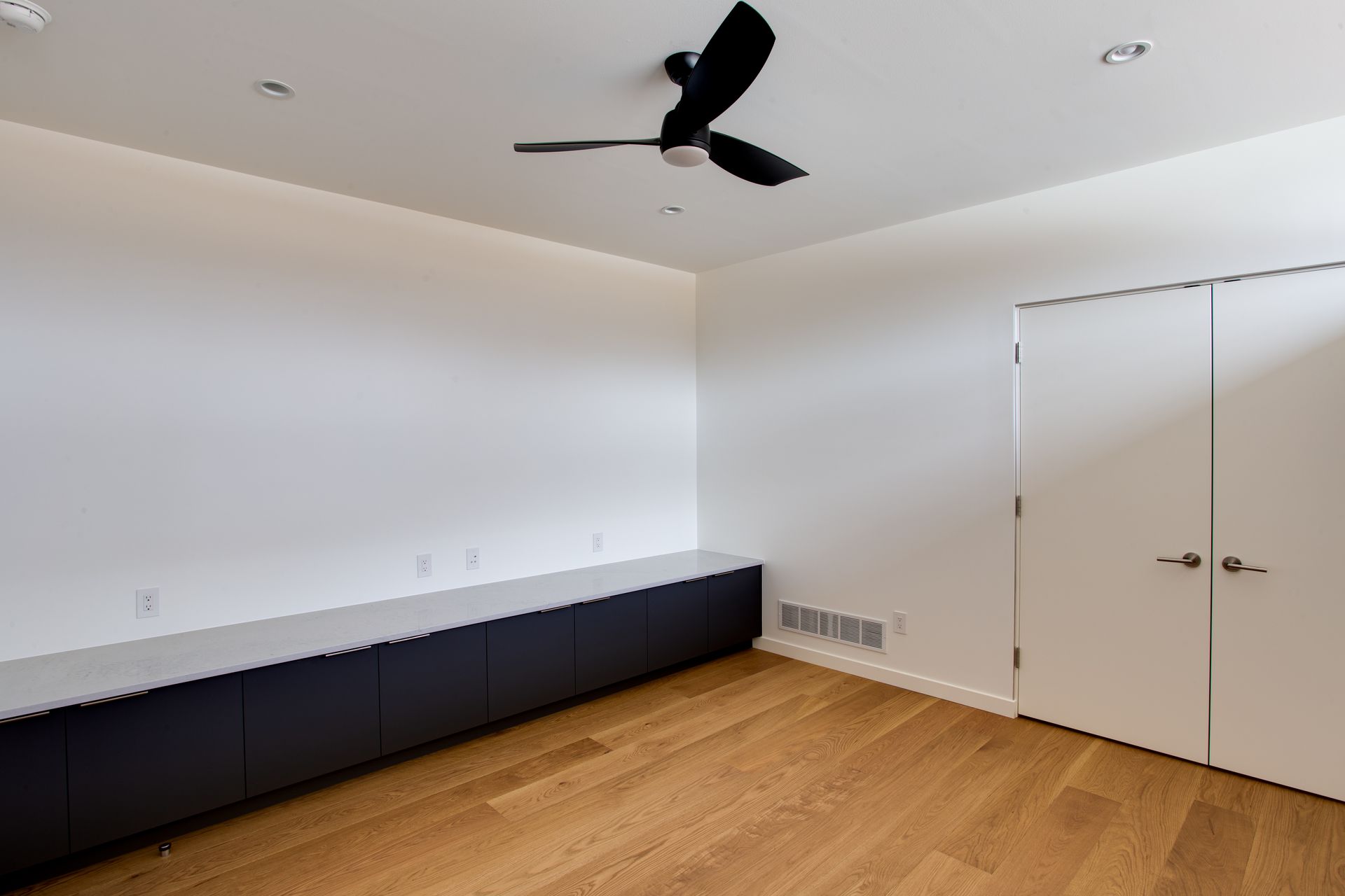 Empty modern bedroom with white walls, wood floor, black ceiling fan, and built-in dark storage cabinets.