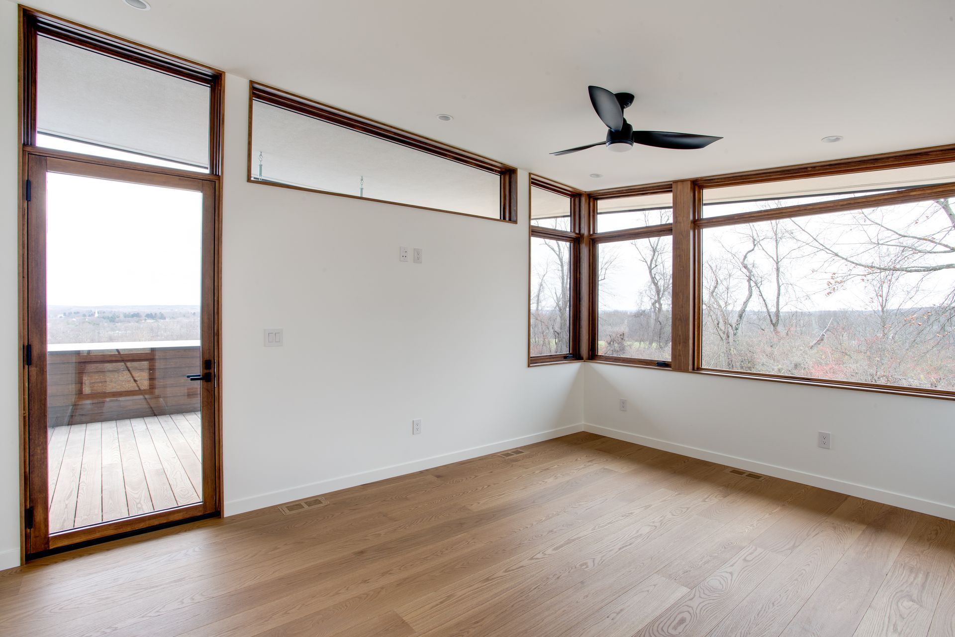Empty room with light wood floors, white walls, large windows, and a ceiling fan.
