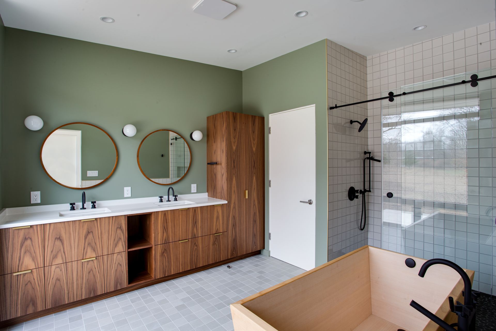Modern bathroom with wood vanity, round mirrors, glass shower, and bathtub under a skylight.
