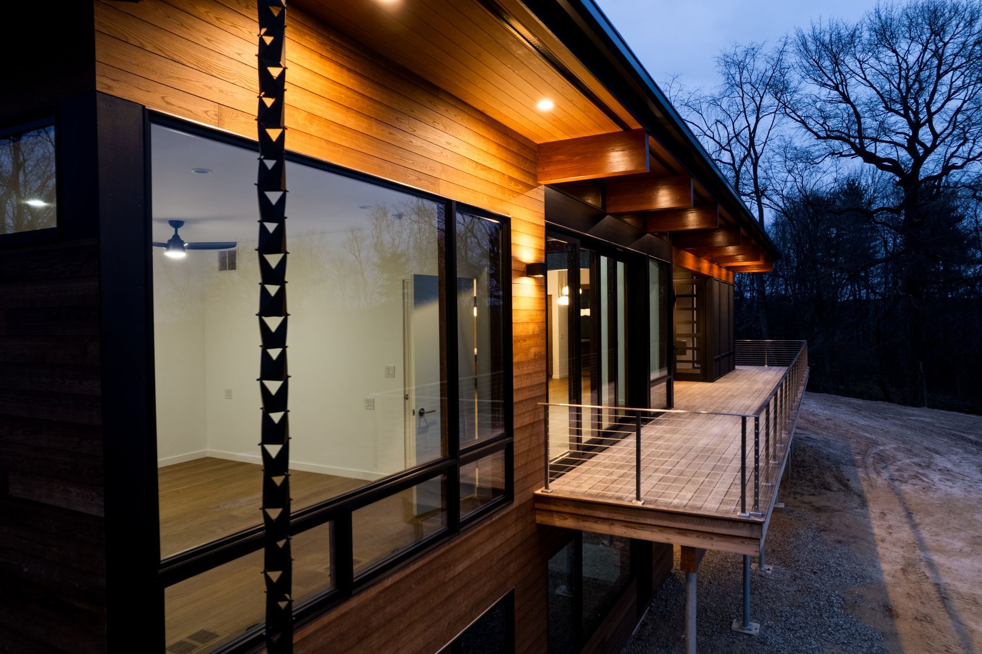 Modern glass building with warm lights and a wooden deck at dusk, beside a gravel path and trees