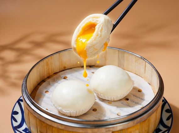 Chopsticks lifting a soft-boiled egg over a bamboo steamer with two eggs inside, yolk dripping.