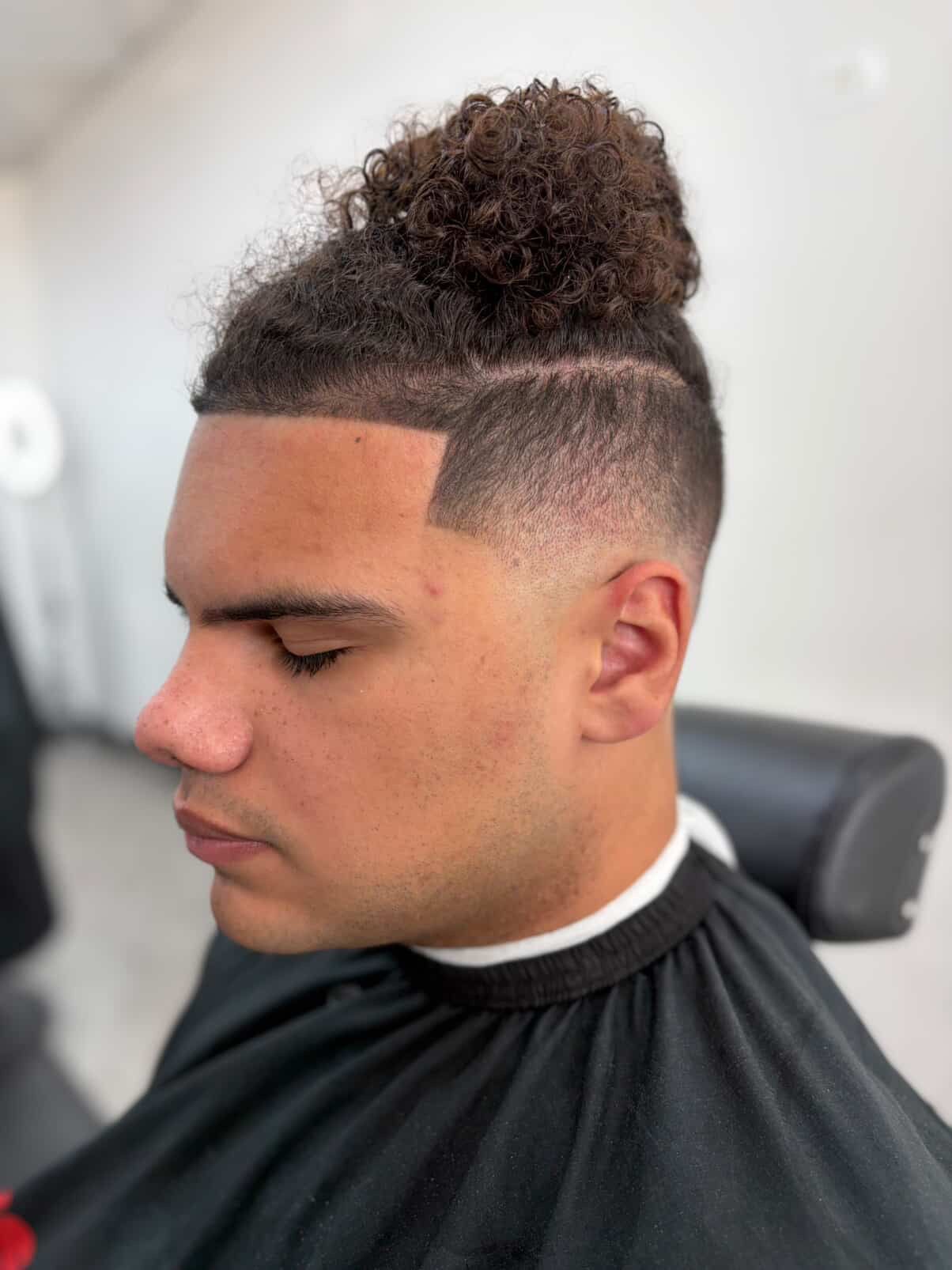 A man with a high-fade haircut and a curly hair bun, viewed from the side while sitting in a salon chair.