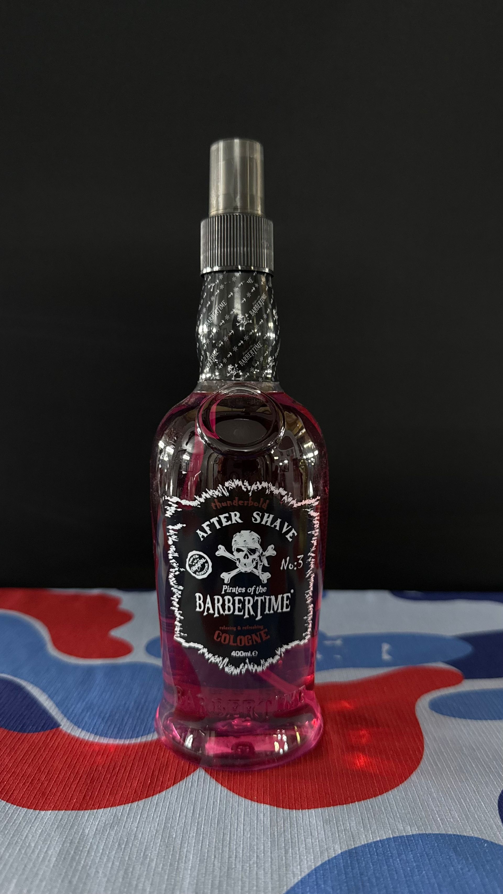 A bottle of Barber Time aftershave with pink liquid, featuring a skull and crossbones logo, standing on a colorful surface.