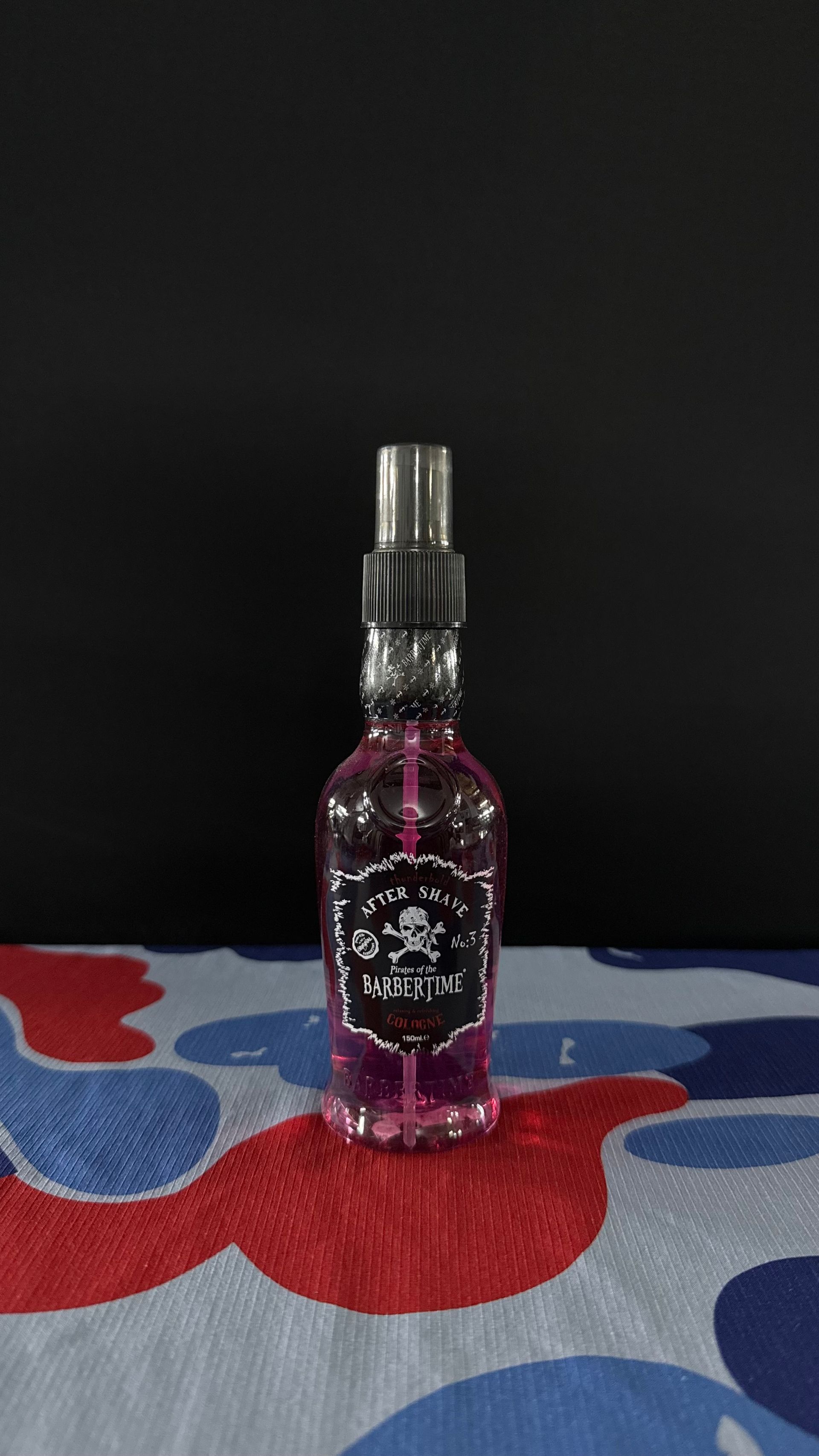A small spray bottle with pink liquid and a skull-and-crossbones label, set on a blue and red camouflage patterned cloth.