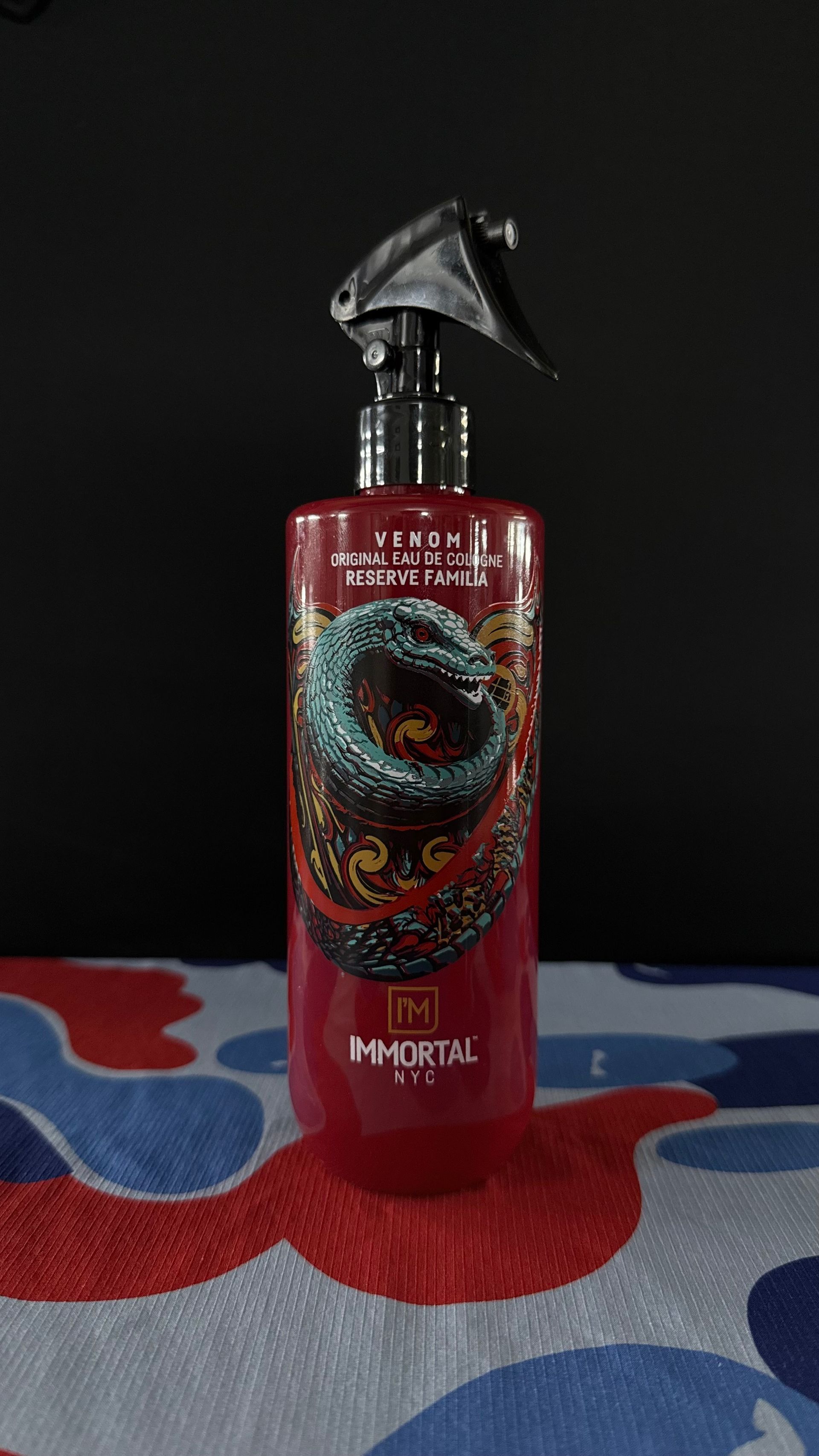 A red spray bottle of Immortal NYC hair styling product featuring a stylized serpent logo on a camo-patterned surface.