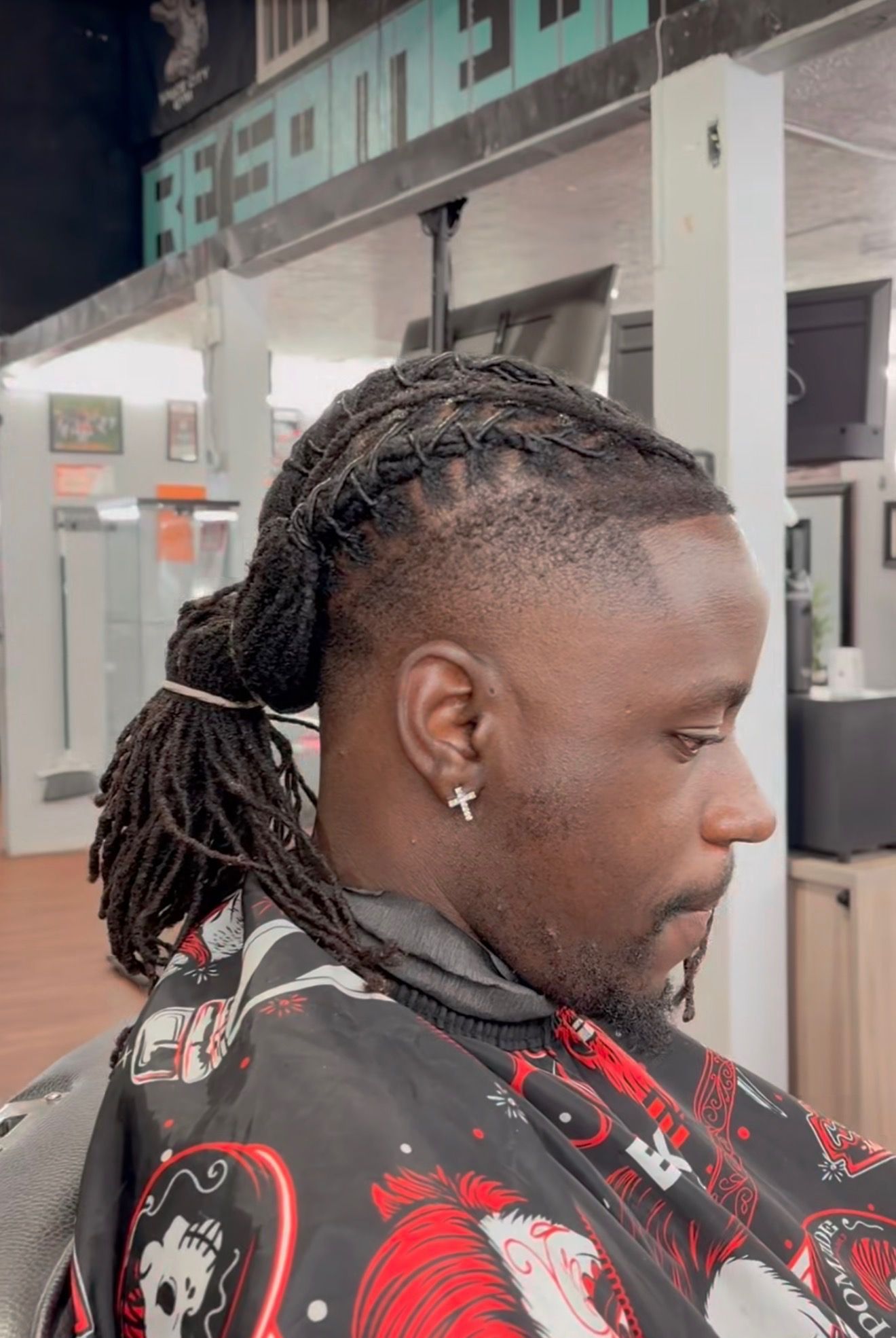 A side profile of a person in a barbershop chair with a low fade haircut and locs gathered into a ponytail.