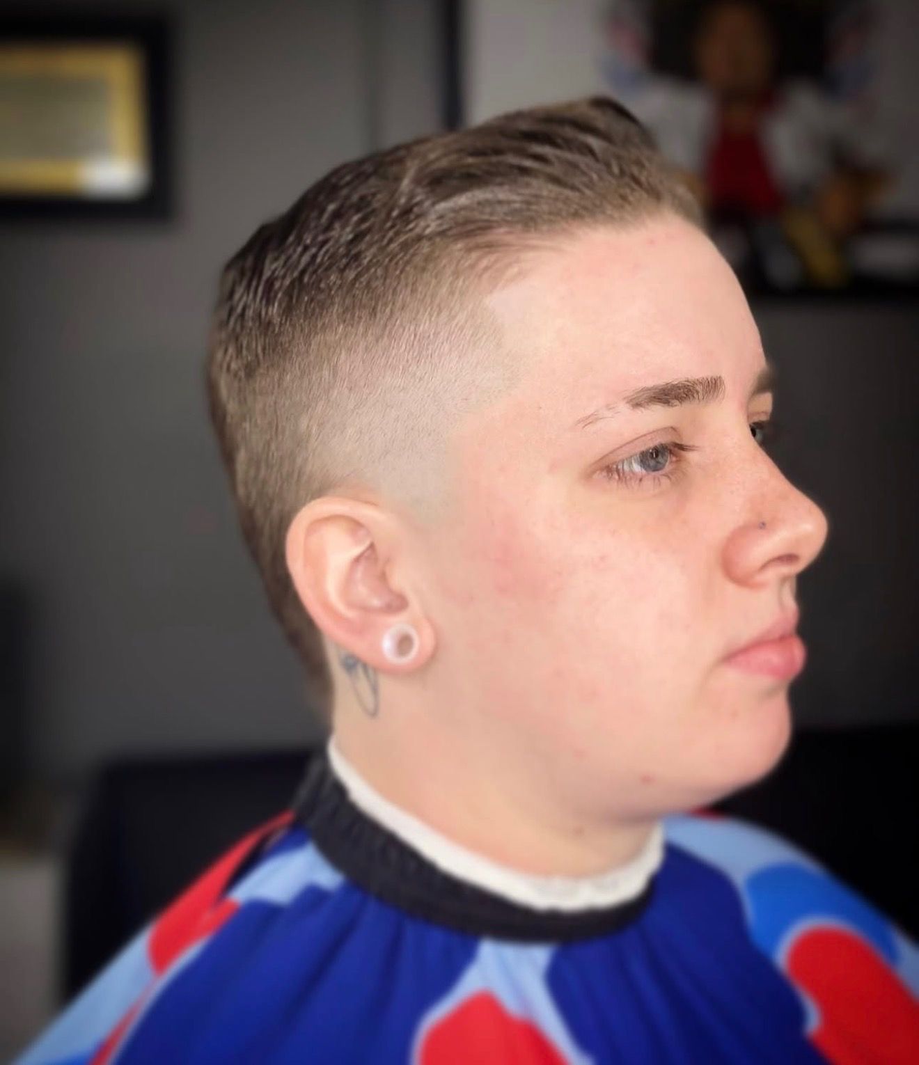 A person with a short, faded haircut and a small ear gauge, wearing a barber’s cape in a salon setting.
