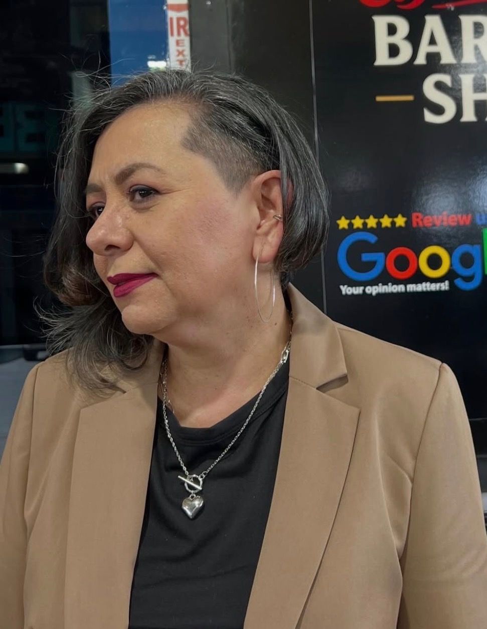 A person with a side-shaved haircut wearing a tan blazer and black top, standing in front of a Google review sign.