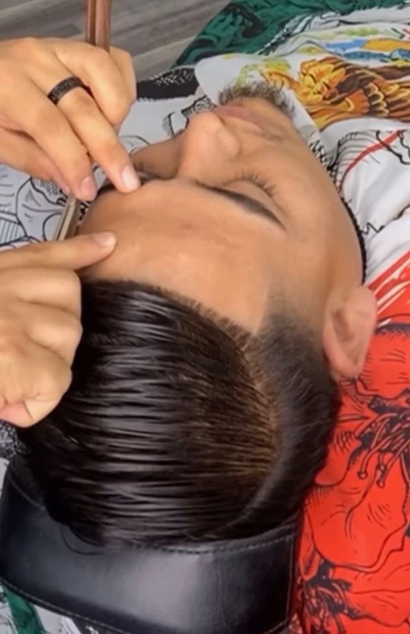 A barber uses a straight razor to groom a client's eyebrow while the client lies back in a chair.
