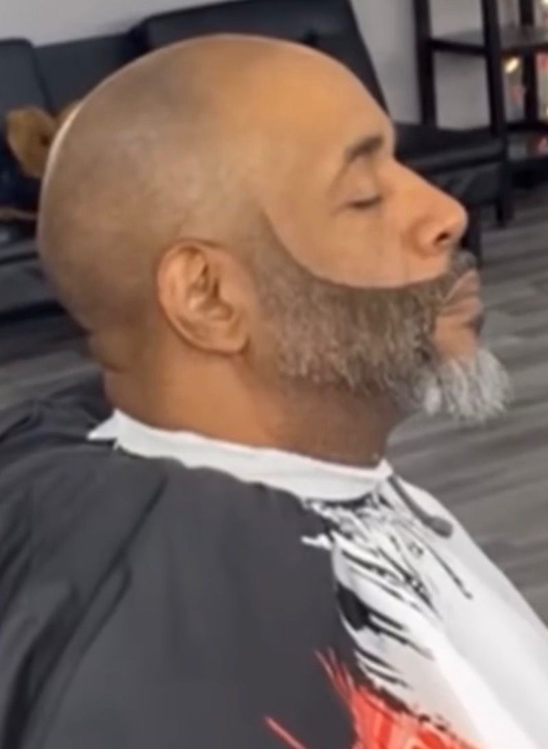 A profile view of a person wearing a barber's cape, featuring a very clean, sharp beard trim and faded neckline.