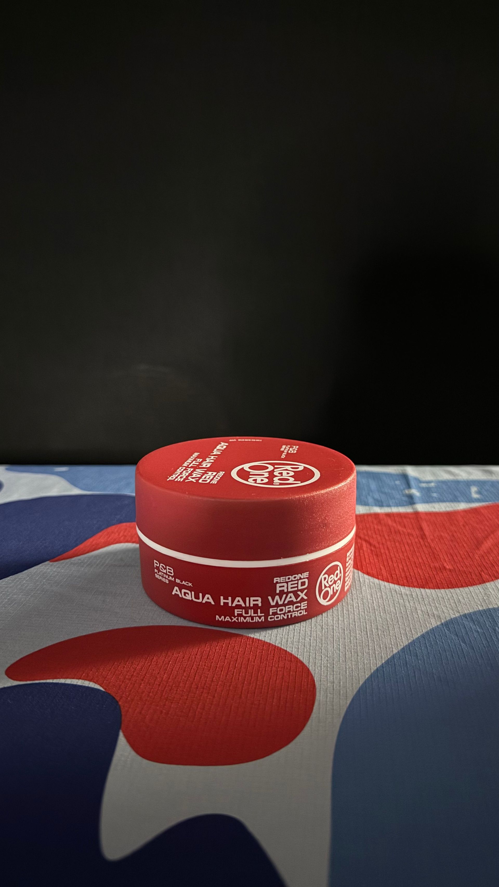 A small, round red container with white text and logos sits on a surface featuring a blue, red, and white camouflage print.