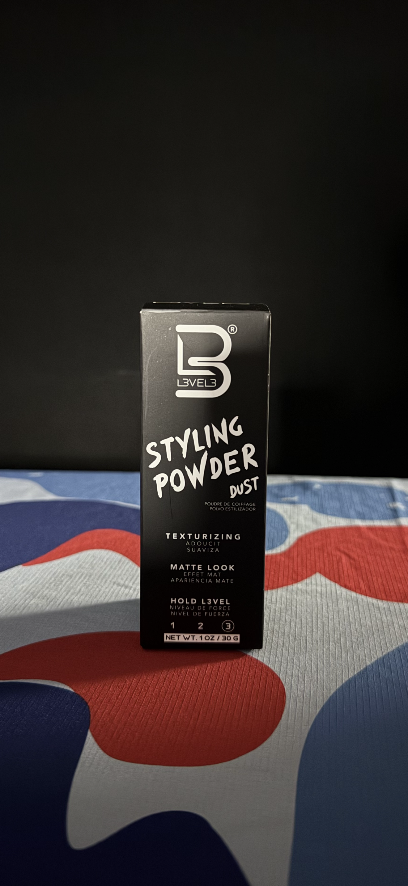 A black bottle of styling powder with white text, standing on a red, white, and blue camouflage patterned surface.