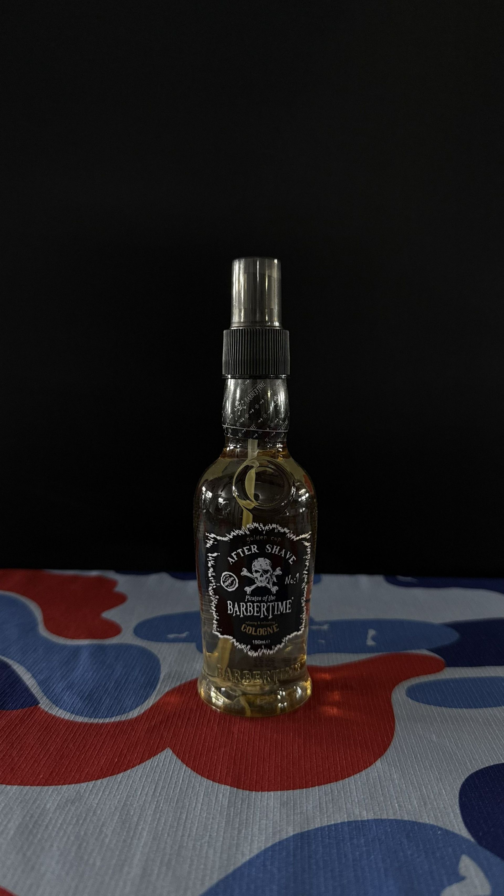 A small, clear glass spray bottle with a black label featuring a skull icon, centered on a red, white, and blue surface.