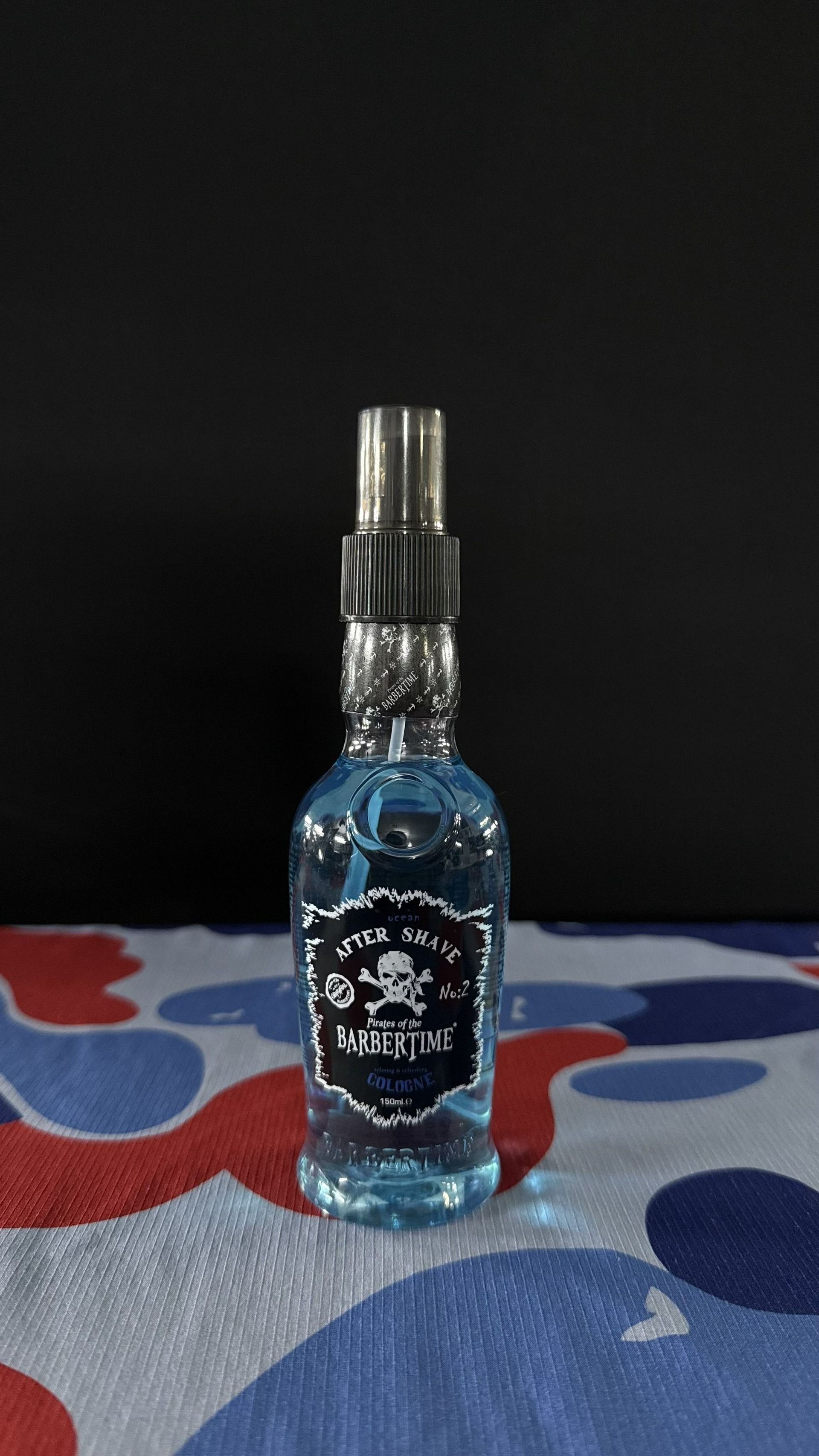 A small, clear glass bottle filled with blue liquid featuring a black skull-and-crossbones label, set on a camo-print fabric.
