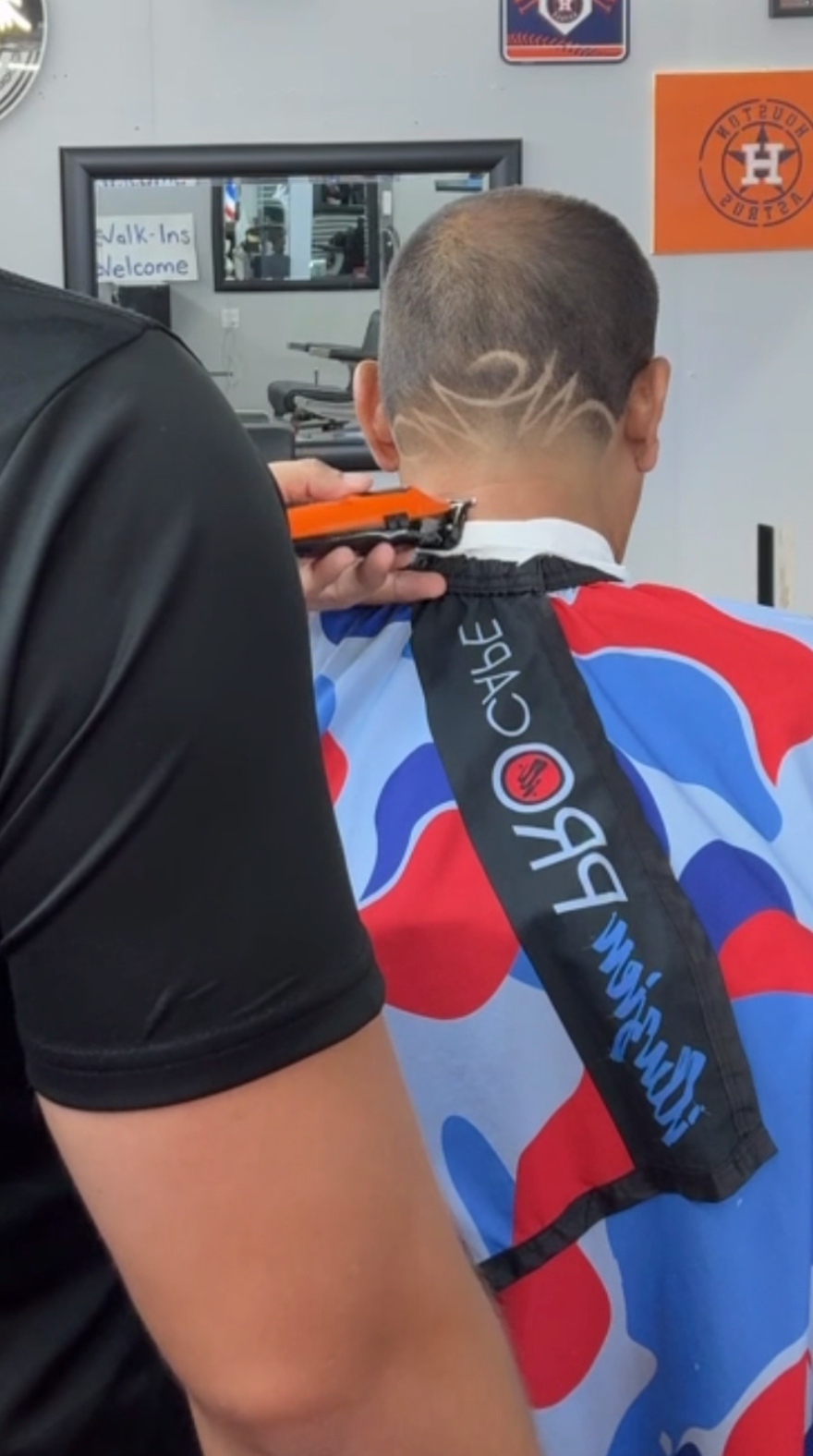 A barber uses clippers to etch a swirling tribal-style design into the back of a person's shaved head in a salon.