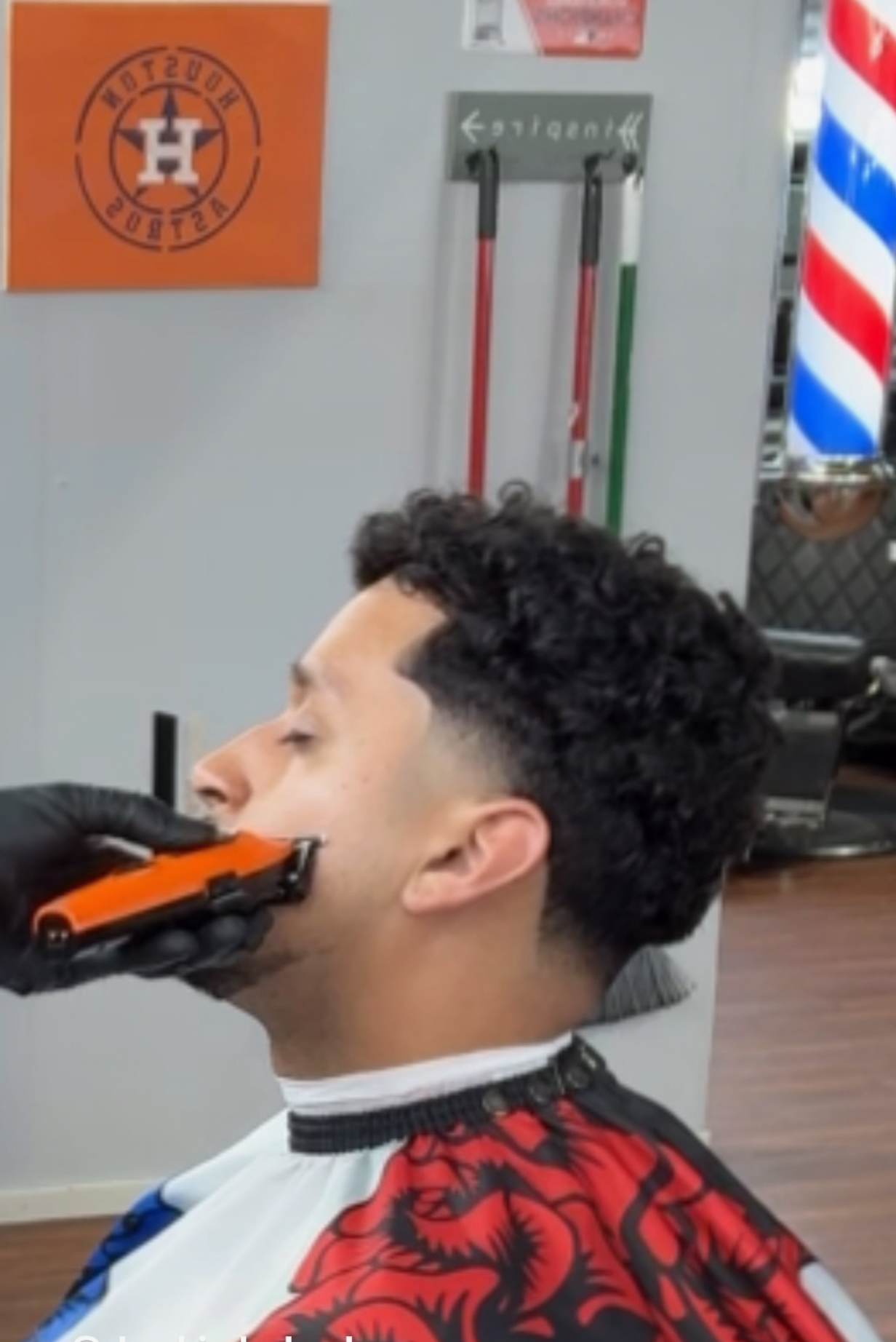 A barber in black gloves uses an orange trimmer to groom a person’s beard in a barbershop.