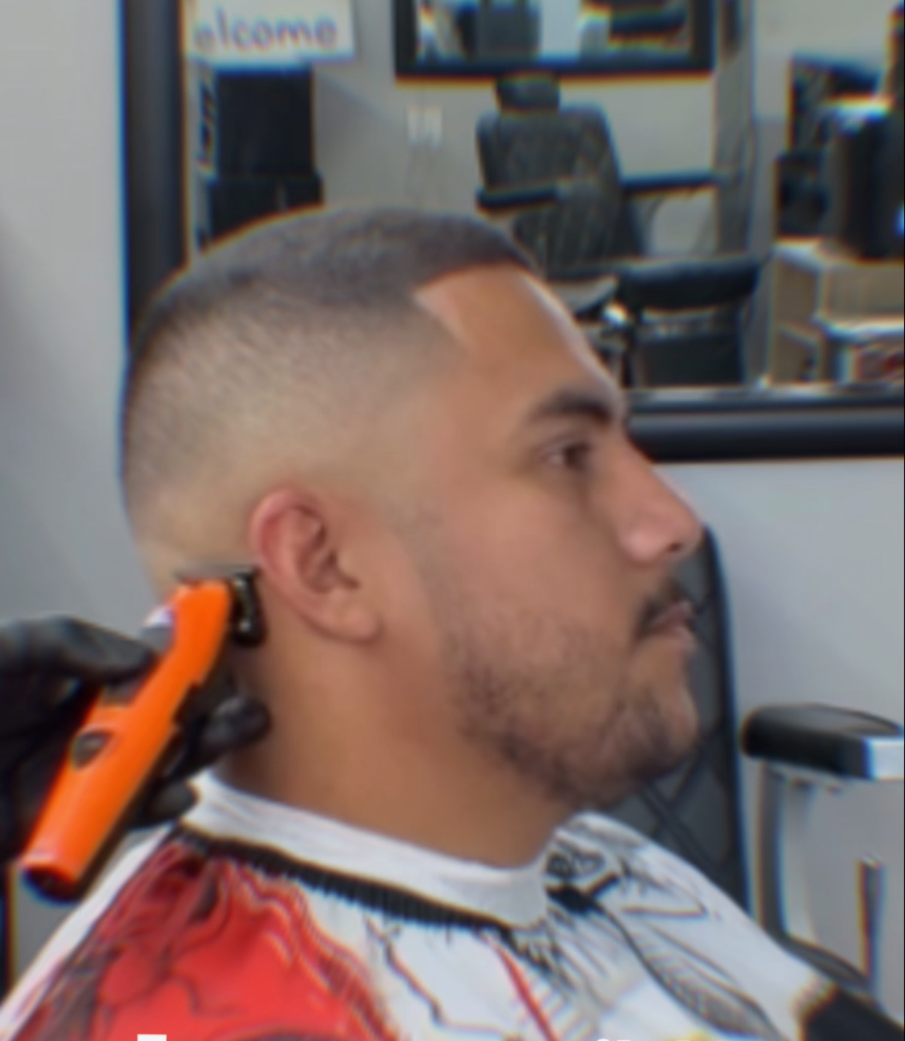 A barber in black gloves uses an orange trimmer to cut hair on the side of a person’s head in a barbershop.