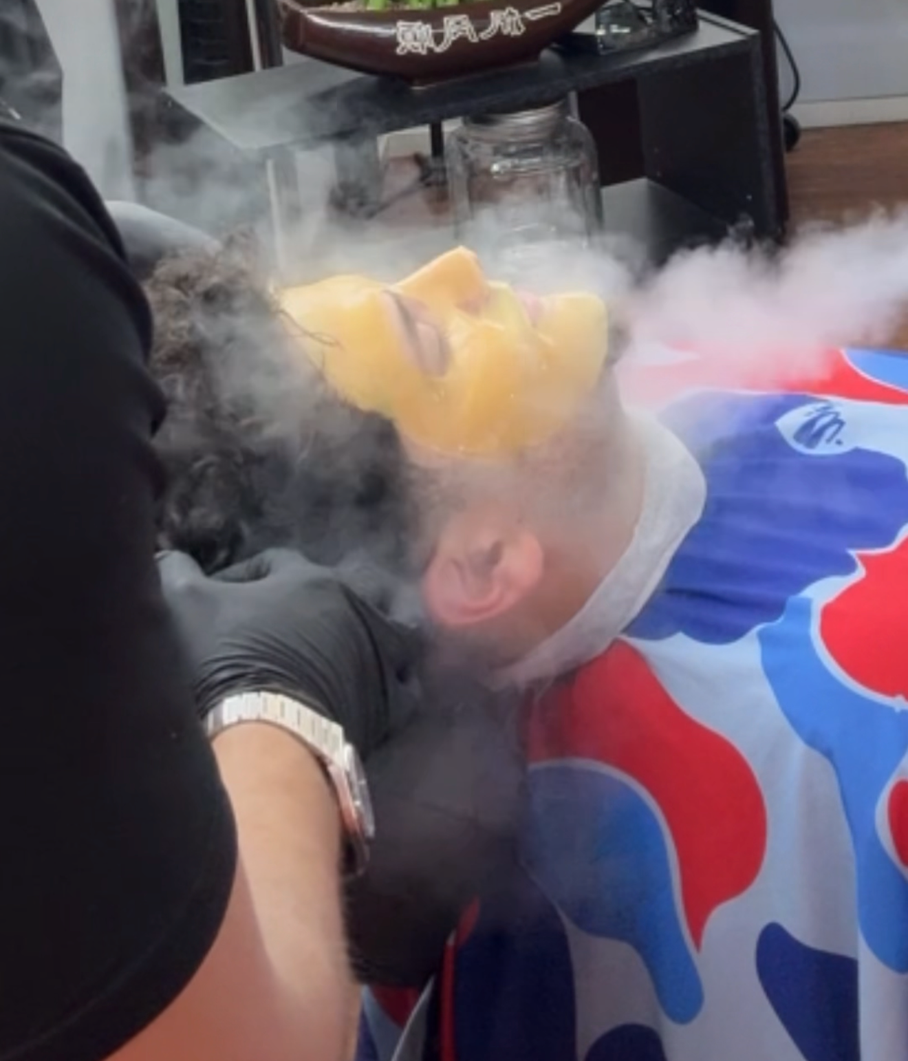 A person lies in a barber chair receiving a facial treatment with a yellow mask while steam surrounds their face.