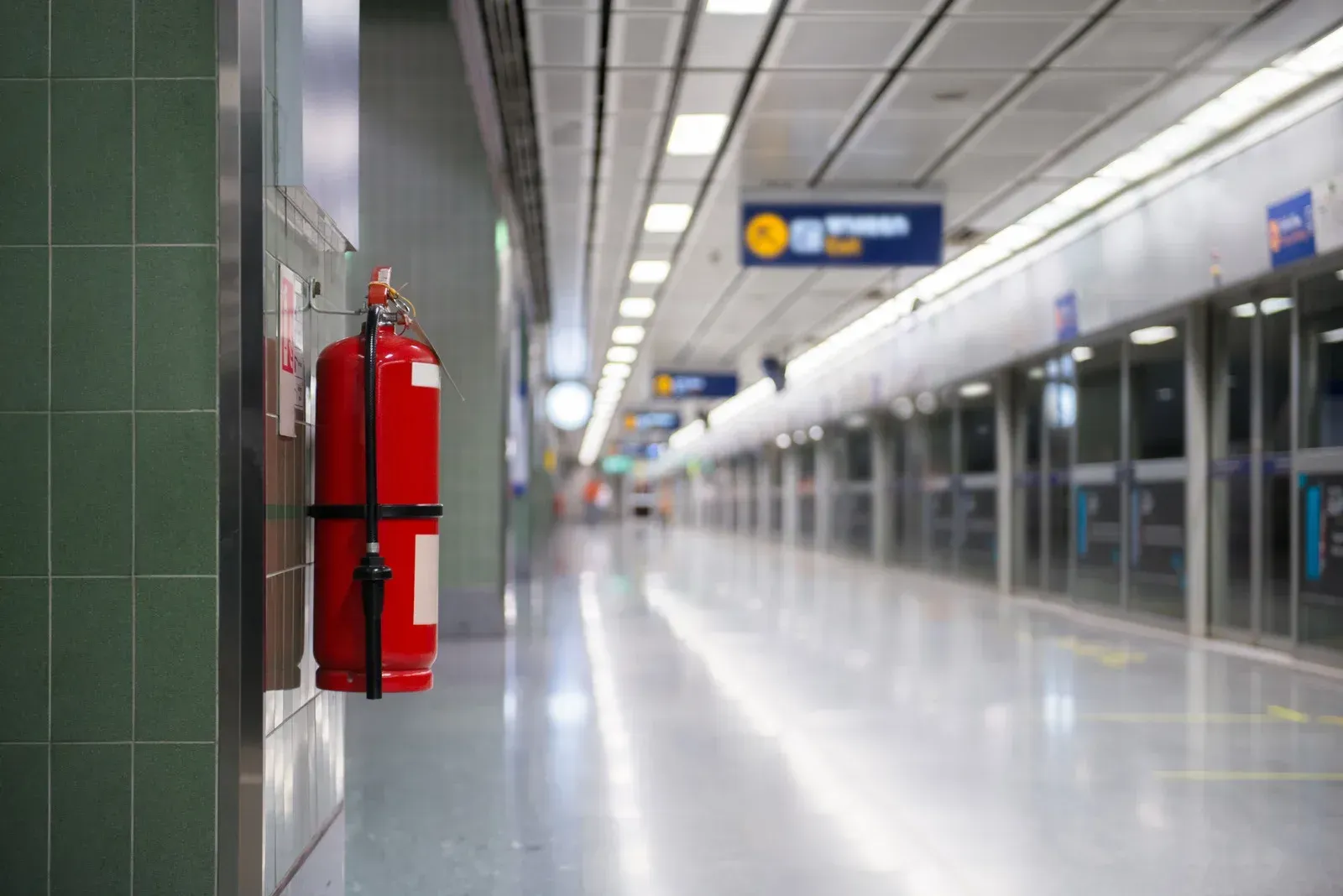 Red fire extinguisher mounted on a green wall in a brightly lit subway station.