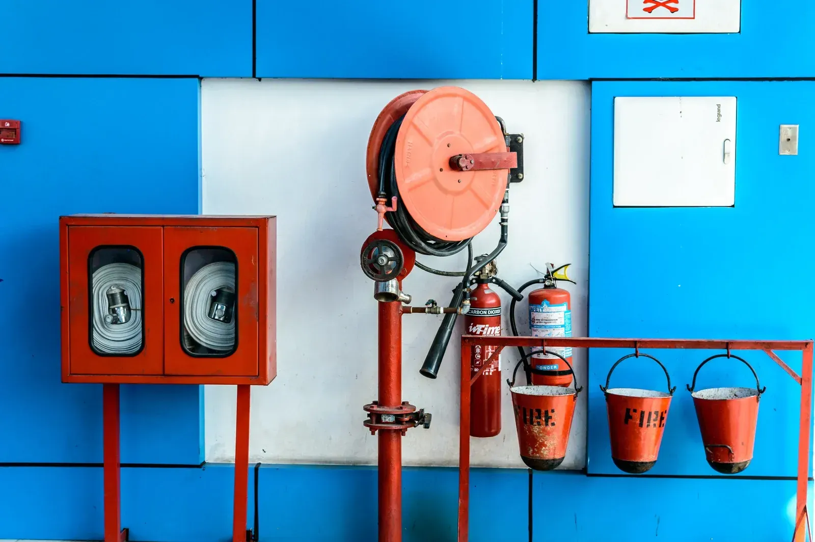 Fire safety equipment: red fire hose reel, extinguishers, buckets, and cabinet on blue and white wall.