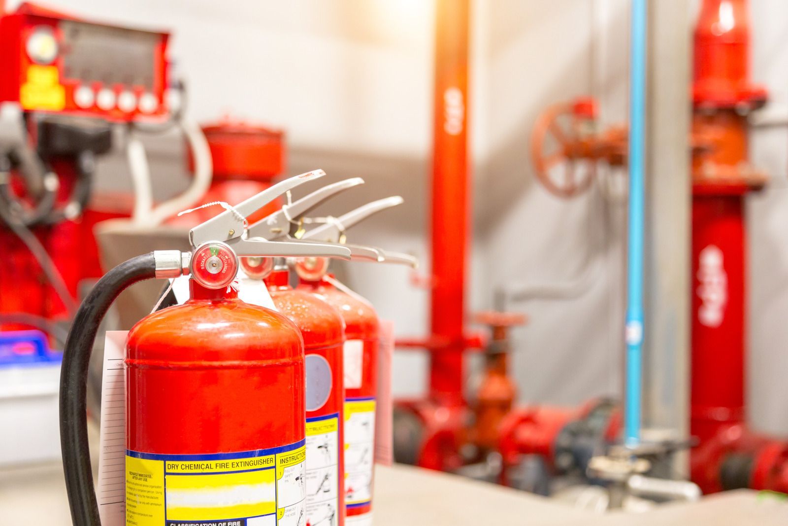 Three red fire extinguishers in front of red pipes and valves, indoor setting.