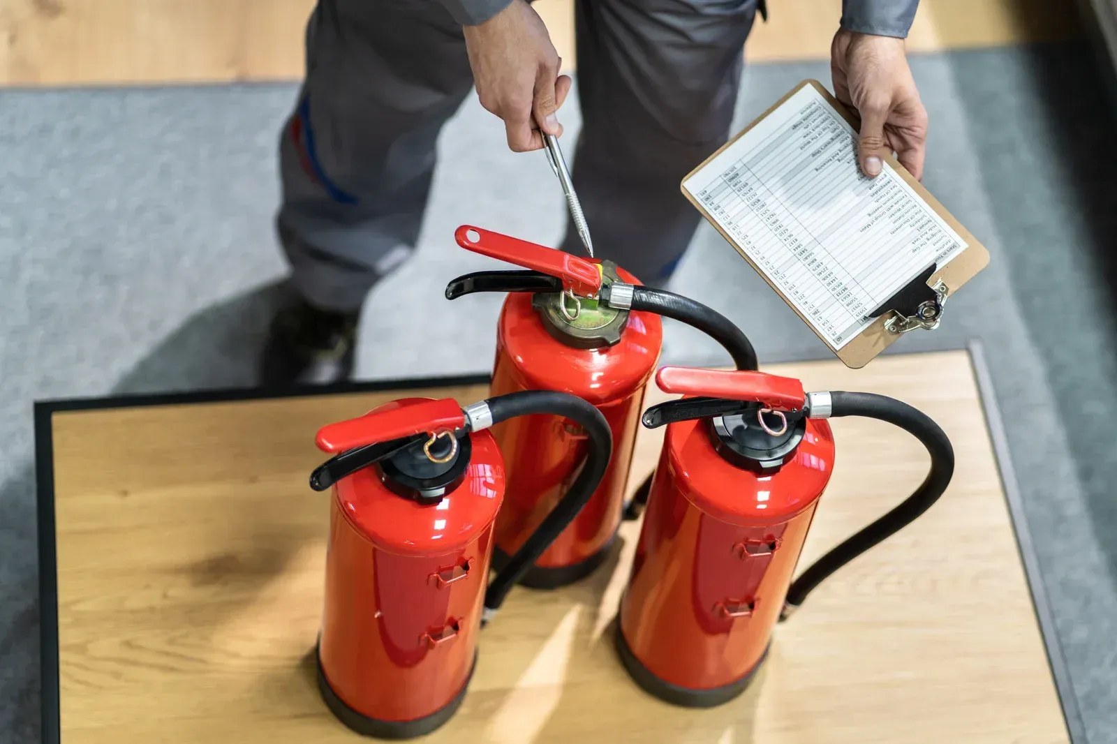 Person inspecting fire extinguishers on a table, holding a clipboard and tool.