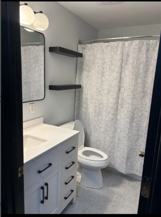 Small bathroom with white vanity, toilet, gray shower curtain, and wall shelves.