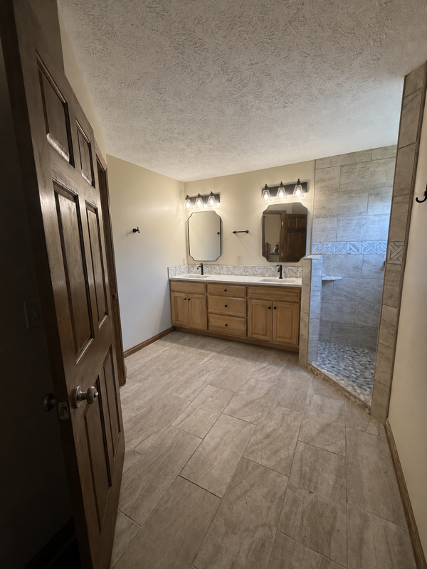 Spacious bathroom with double vanity, mirrors, tiled shower, and dark wood door on the left.