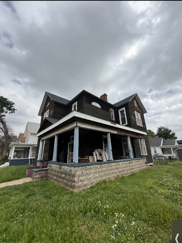 Large dark house with a wraparound porch and stone foundation, viewed from a grassy yard under cloudy skies