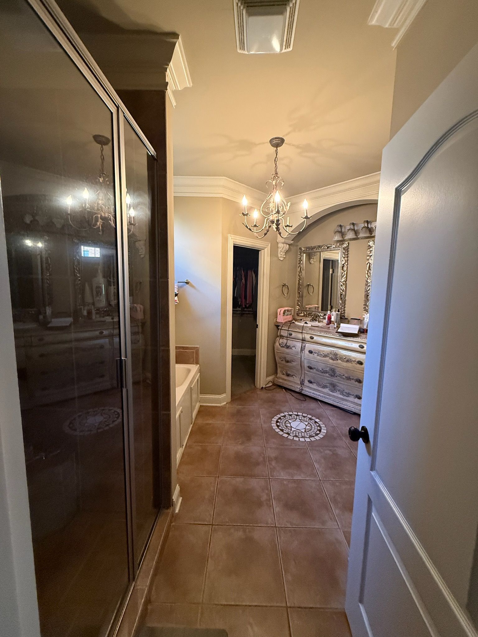 Narrow hallway with tiled floor, chandelier, dresser with mirror, and an open white door in the foreground