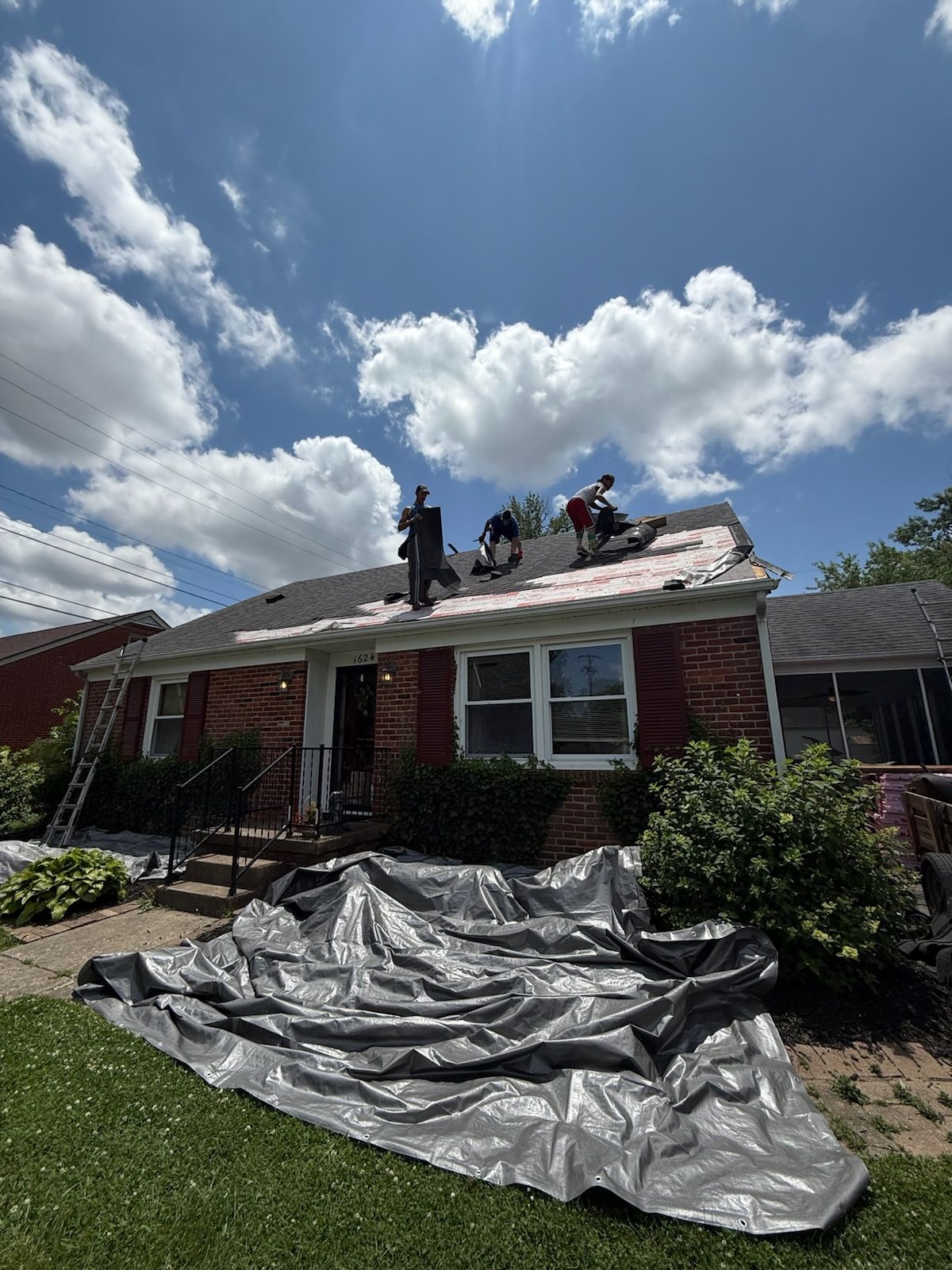 Workers repairing a red house roof with gray tarp on the lawn under a cloudy blue sky
