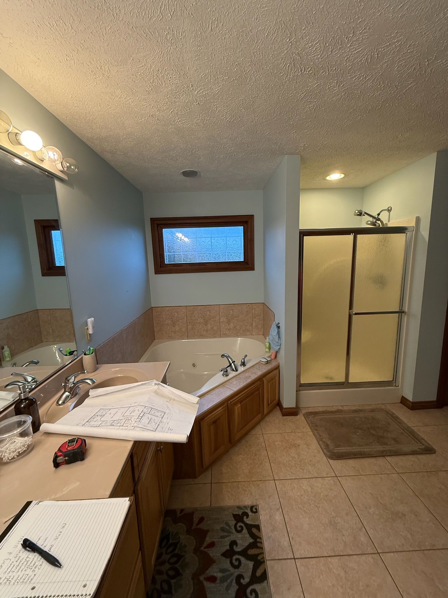 Spacious bathroom with double sink vanity, tiled tub, shower stall, window, and tan flooring.