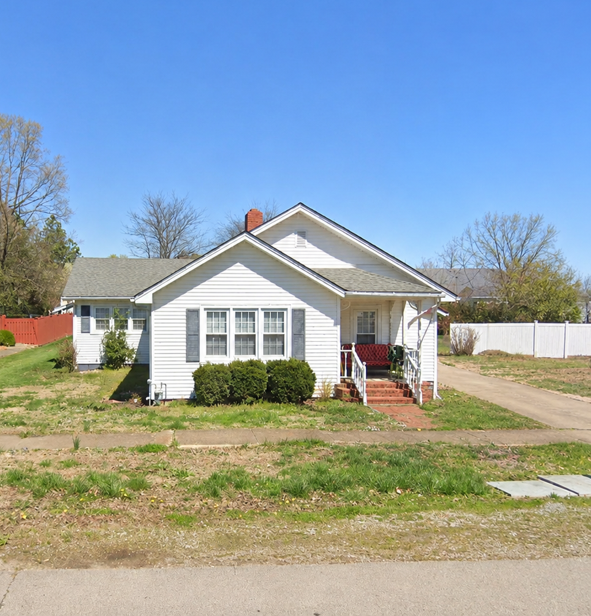 Small white house with a front porch, red steps, and a grassy yard under a clear blue sky