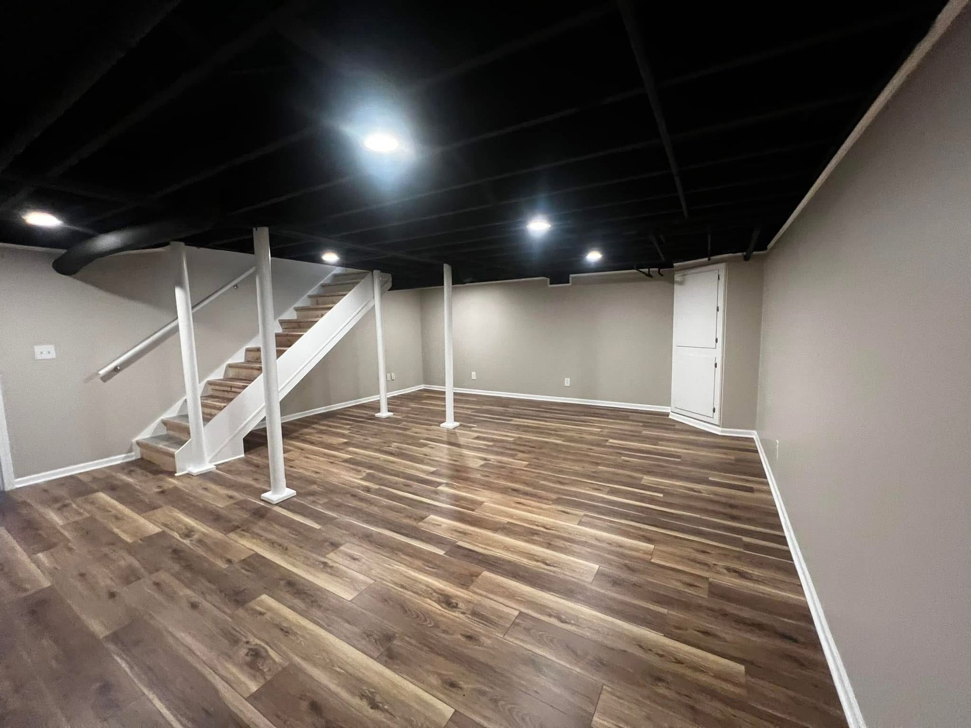Empty basement room with wood floors, white stairs, and black ceiling lights