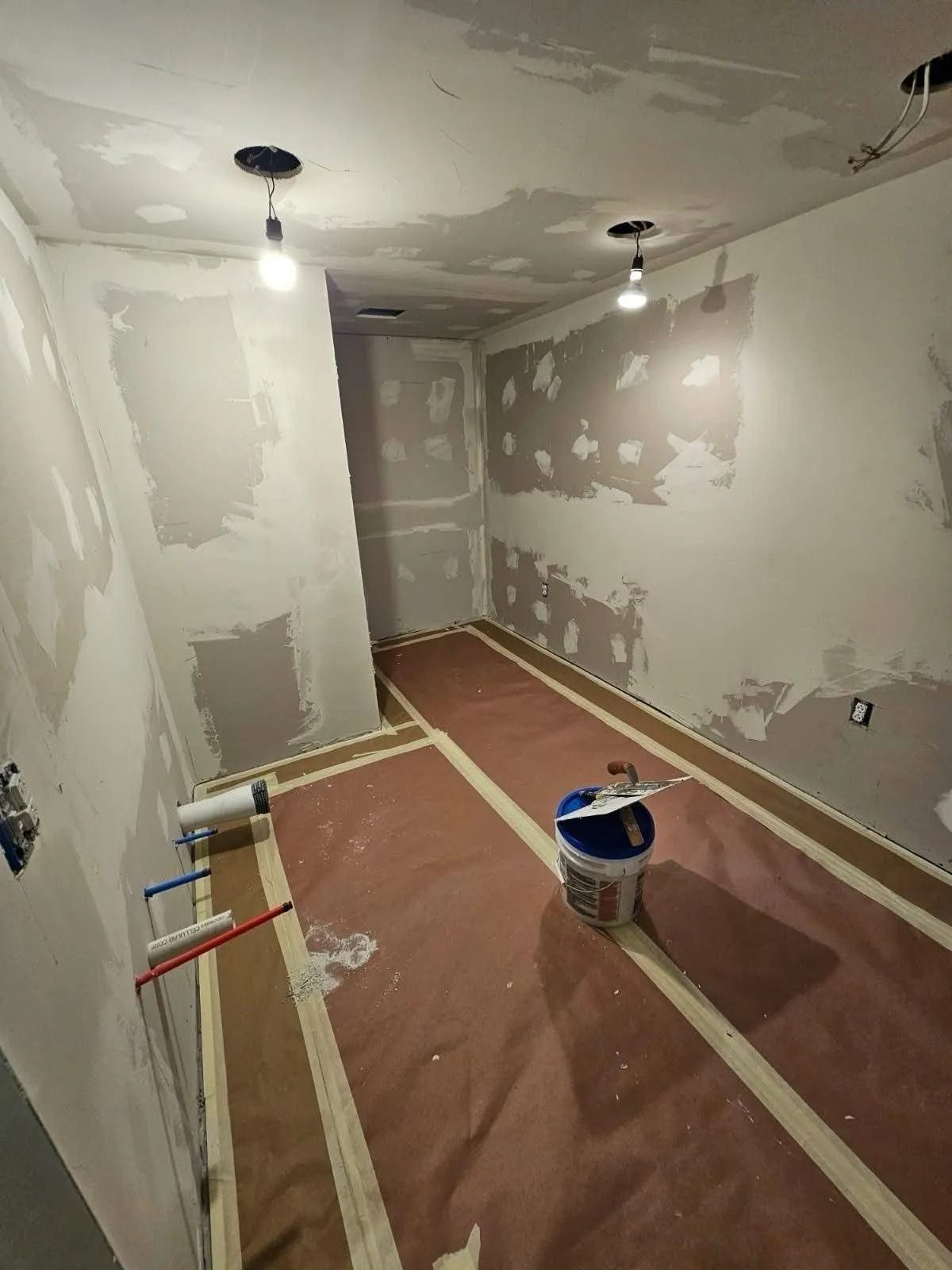 Unfinished room with taped floor, drywall patches, tools, and bare lightbulbs during renovation.
