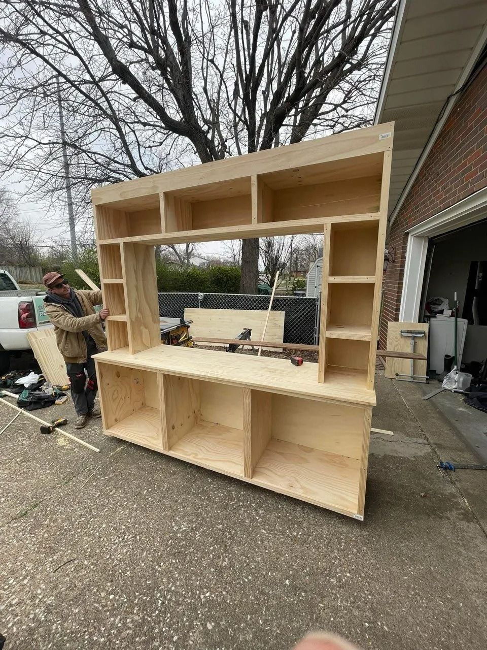 Large unfinished plywood shelving unit outdoors beside a driveway, with a person working nearby.