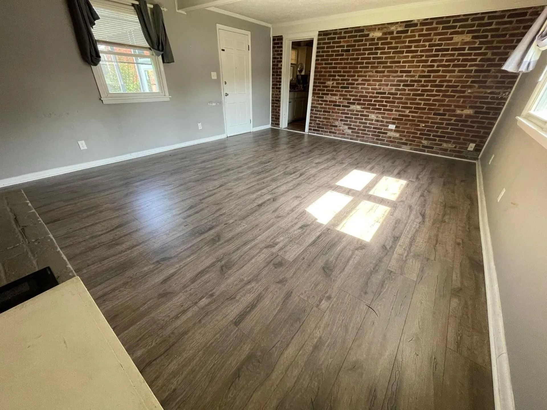 Empty living room with gray wood floors, exposed brick wall, and sunlight from windows.