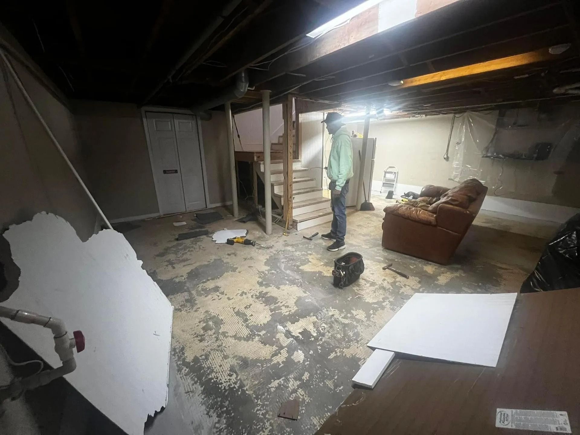 Damaged basement with peeling floor, exposed beams, a couch, and scattered debris after flooding or fire.
