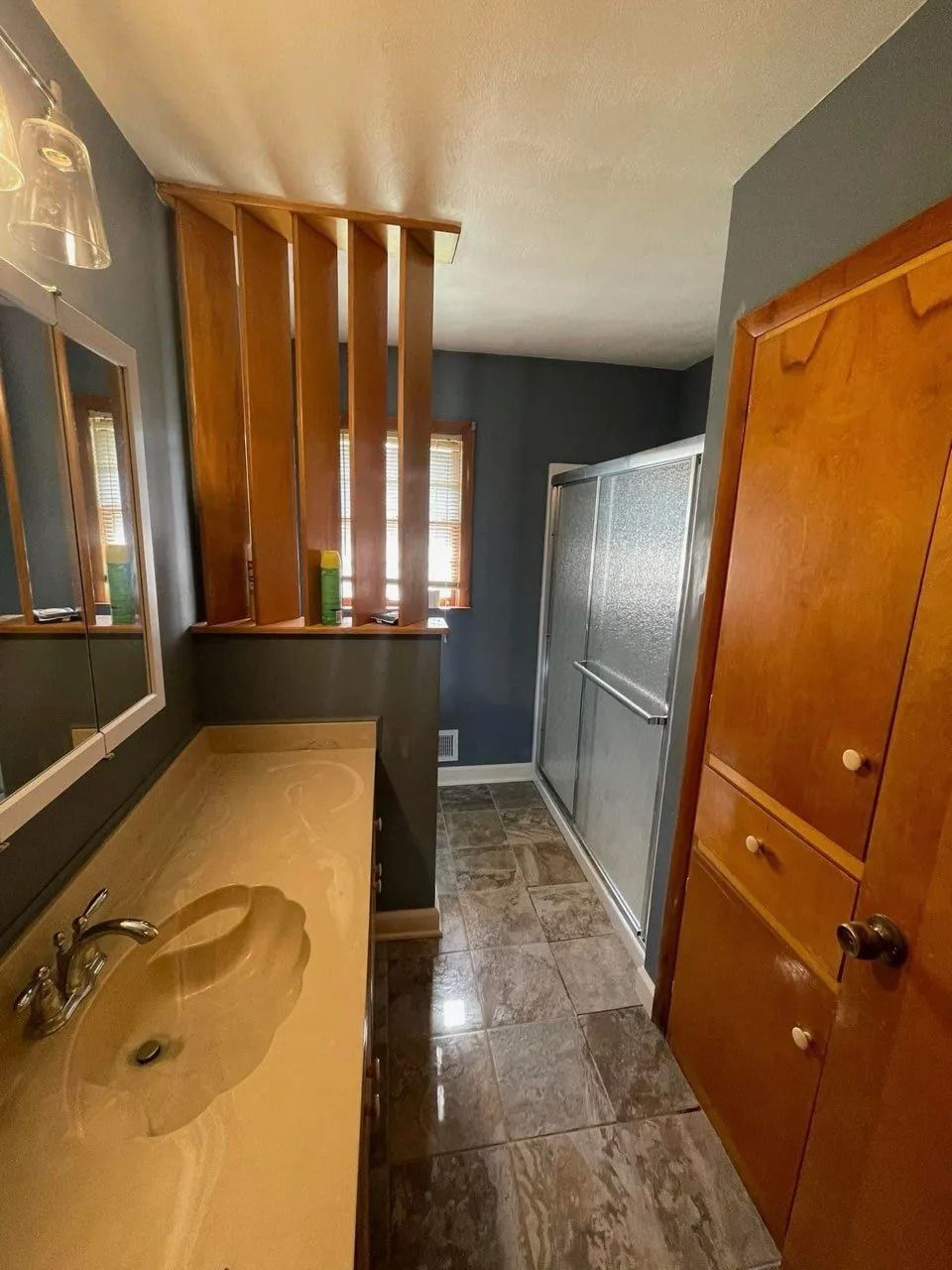 Narrow bathroom with sink, mirror, wooden cabinet, tiled floor, and shower stall beside window blinds