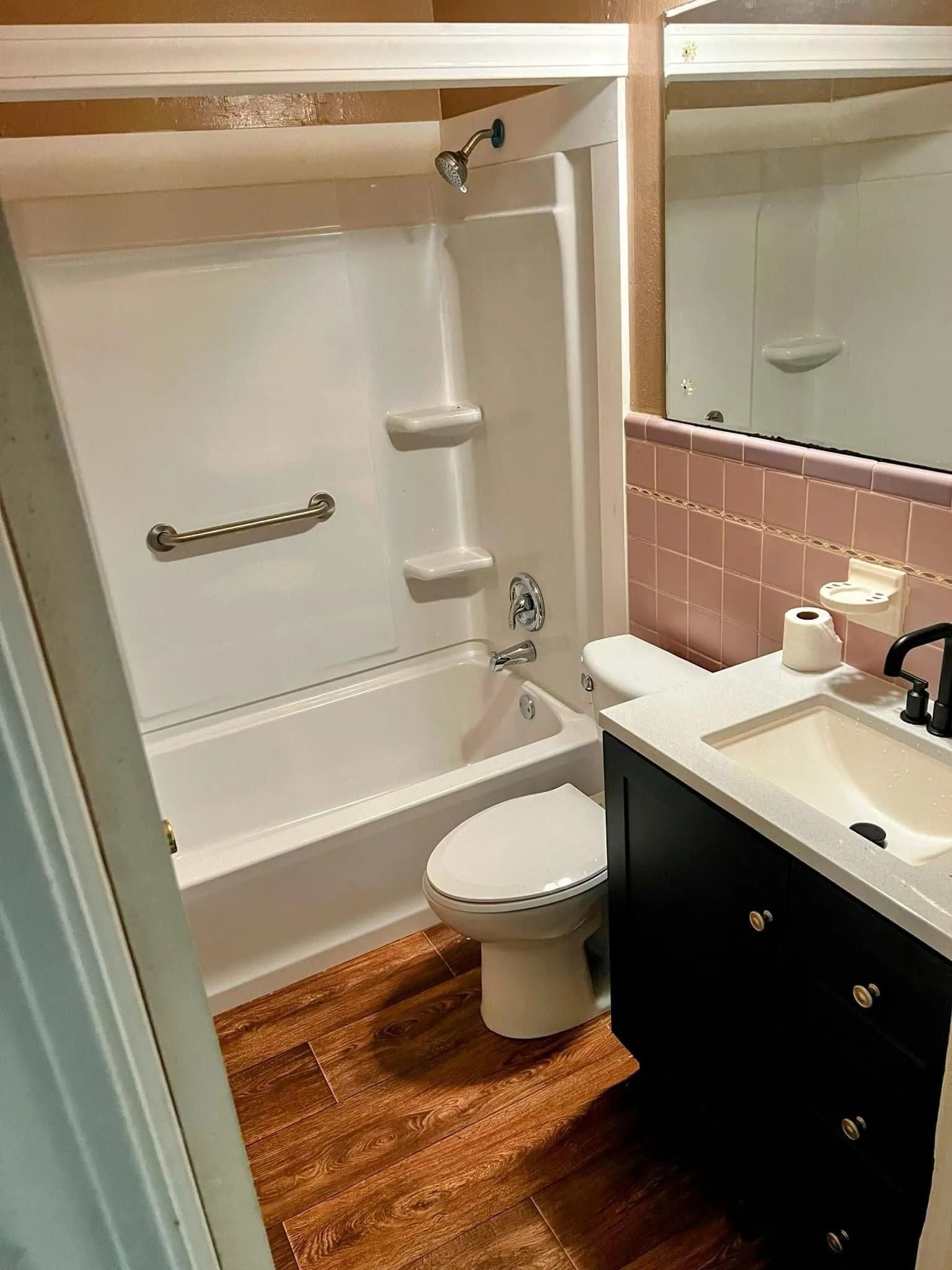 Small bathroom with shower, toilet, sink, black vanity, pink tile, and wood-look floor.