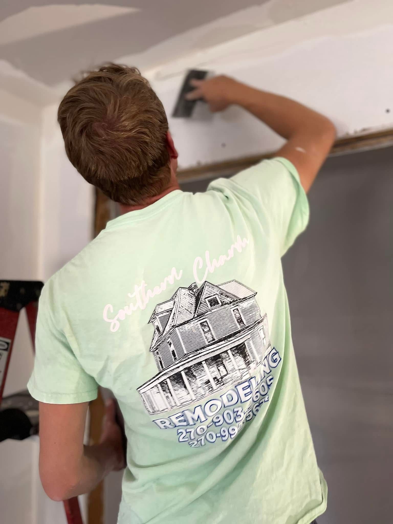Person painting a wall with a roller, wearing a light green T-shirt and facing away from the camera