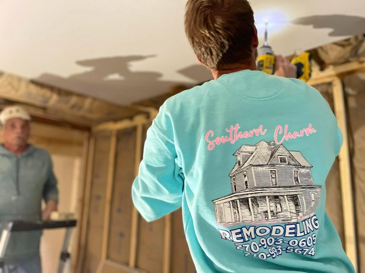Two workers in a room; one wears a turquoise shirt with a house-remodeling logo.