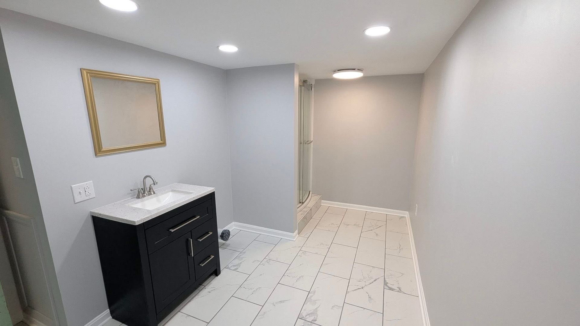 Small bathroom with gray walls, white tile floor, black vanity, and gold-framed mirror.