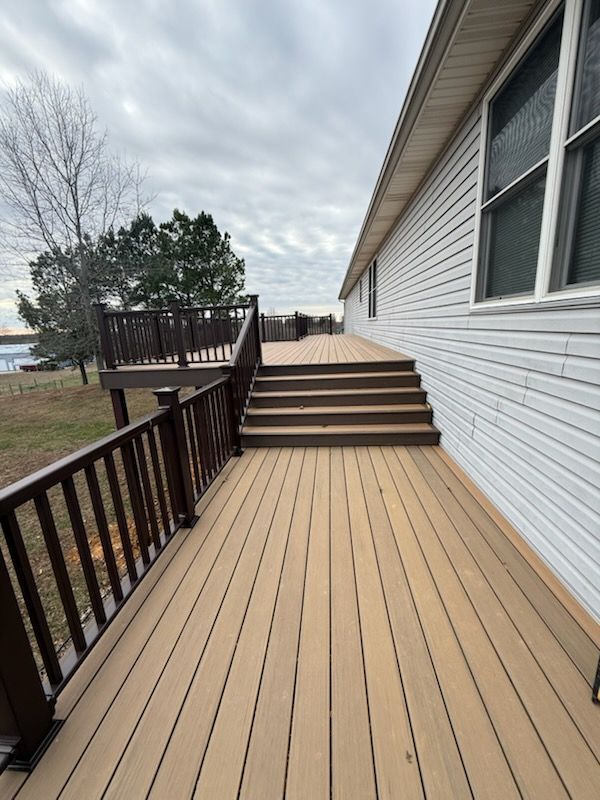 Wooden deck walkway beside a white house, leading up steps to a fenced patio by the water.