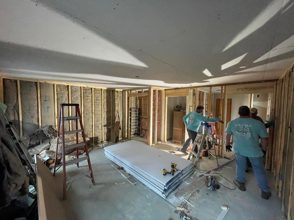 Two workers framing interior walls in a room under renovation, with lumber, ladders, and tools scattered about.
