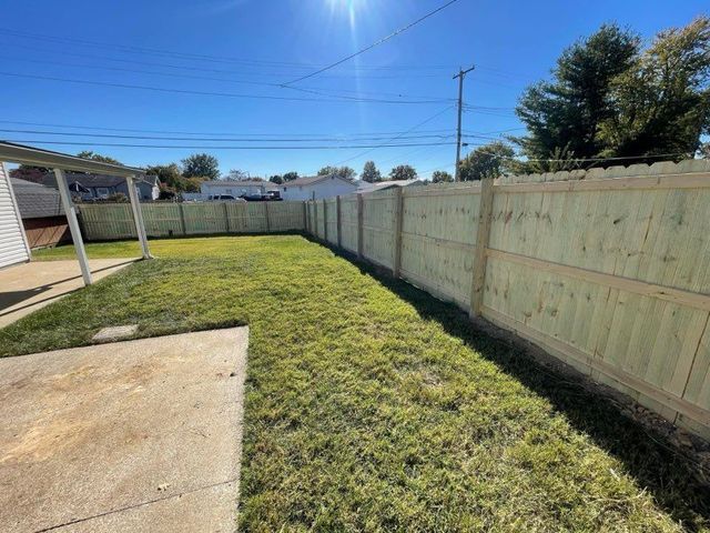 Backyard with green lawn, concrete patio, wooden fence, and a covered patio under a clear blue sky