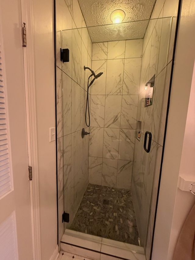 Glass shower stall with marble-look walls, stone tile floor, and a rain showerhead inside a bathroom.